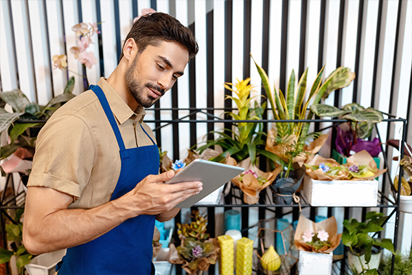 man in a blue apron looking at a tablet and flowers