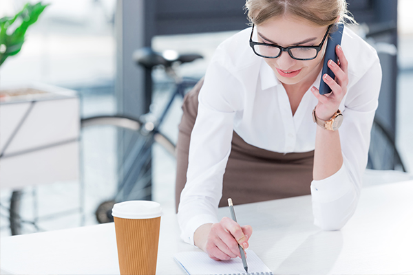 woman in a white blouse on talking on a cellphone while writing in a pad