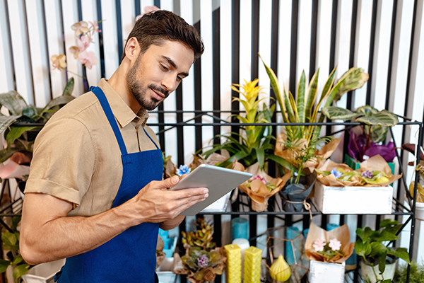 man in a blue apron looking at a tablet and flowers